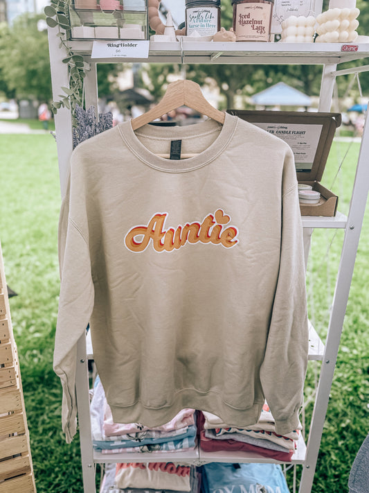 Beige sweatshirt with 'Auntie' text on a hanger against a green outdoor background