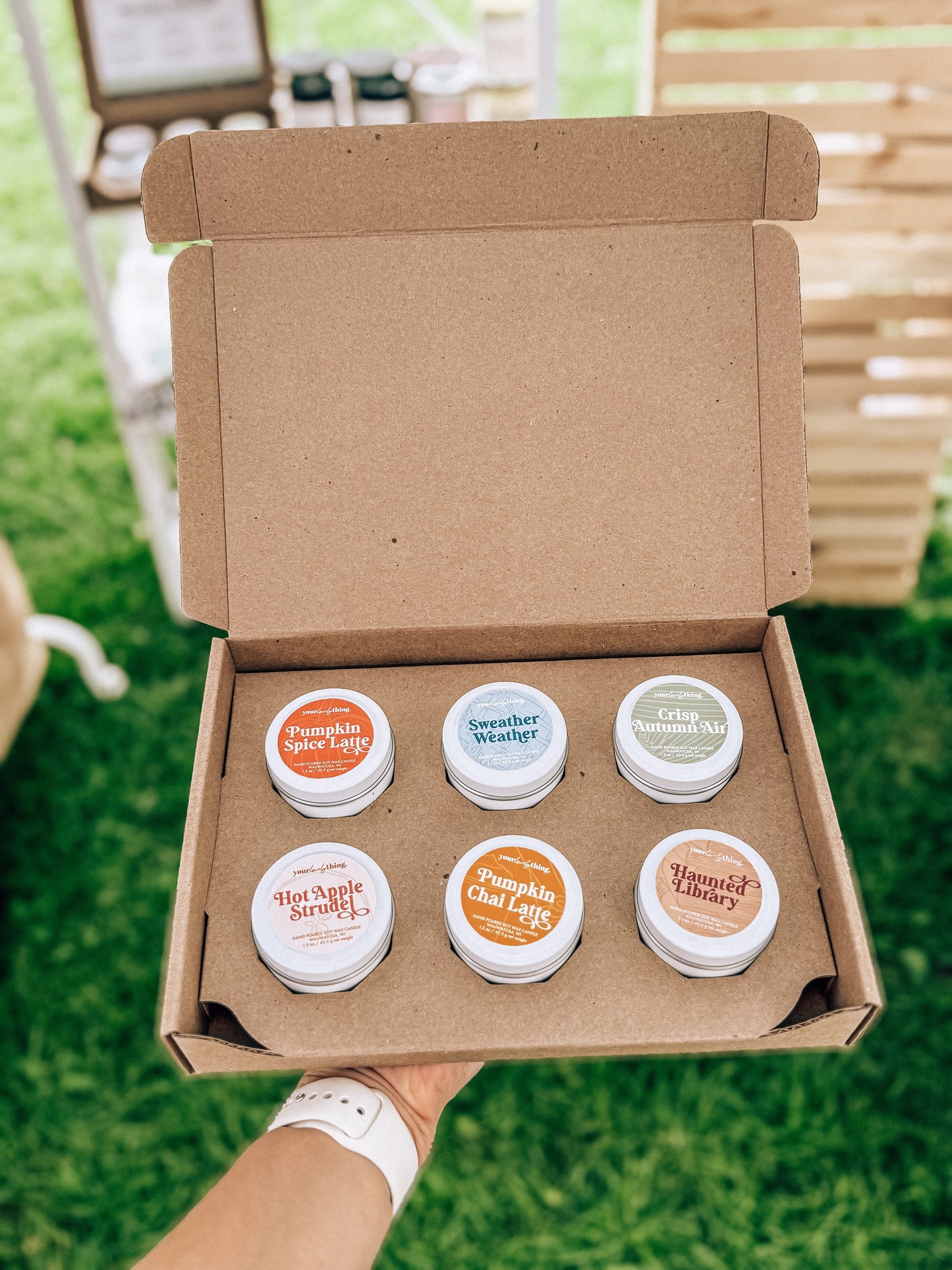 Open cardboard box with six small containers of lip balm on a grassy outdoor background