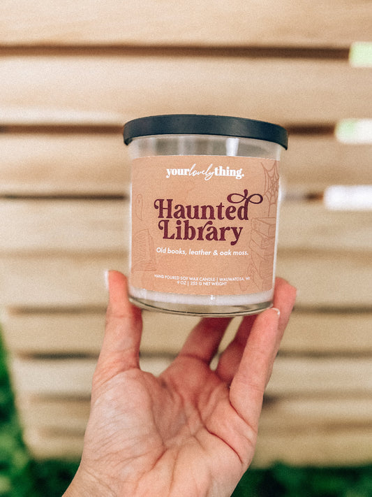Hand holding a candle labeled 'Haunted Library' against a wooden slat background