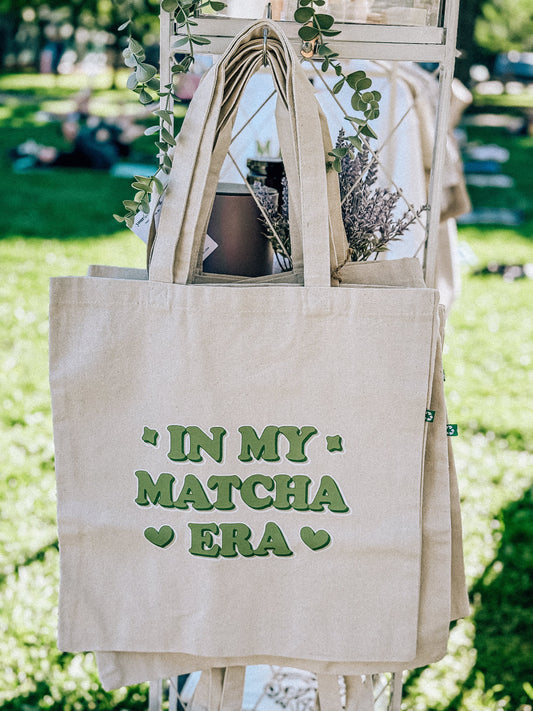 Beige tote bag with 'In my matcha era' text on a blurred outdoor background