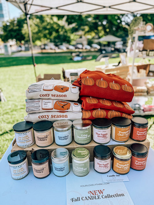 Stack of candles and fabric with 'cozy season' text at an outdoor market.