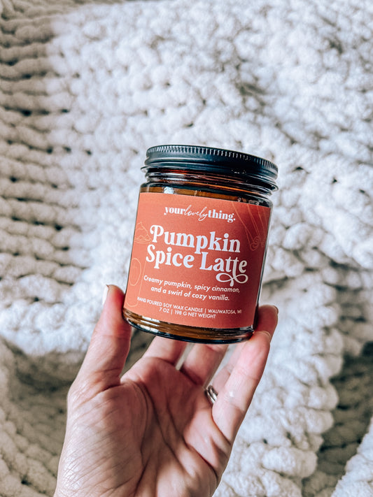 Hand holding a jar of 'Pumpkin Spice Latte' candle with a textured white surface in the background