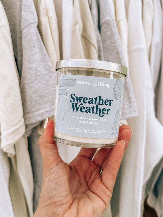 Hand holding a candle labeled 'Sweater Weather' with clothing in the background