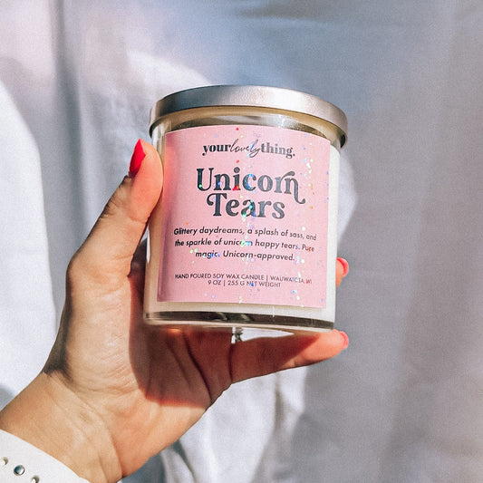 Hand holding a candle labeled 'Unicorn Tears' against a plain background
