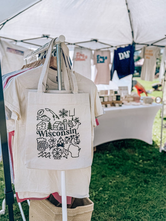 Tote bag with 'Wisconsin' design hanging under a tent at an outdoor event.