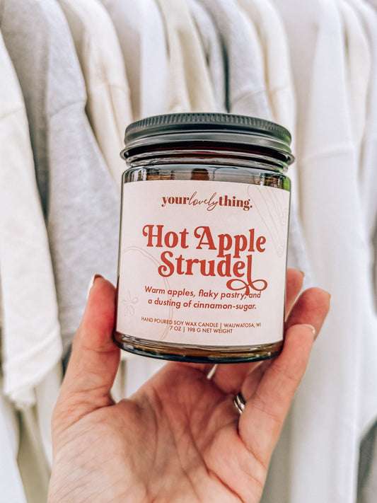 Hand holding a candle labeled 'Hot Apple Strudel' with a neutral background
