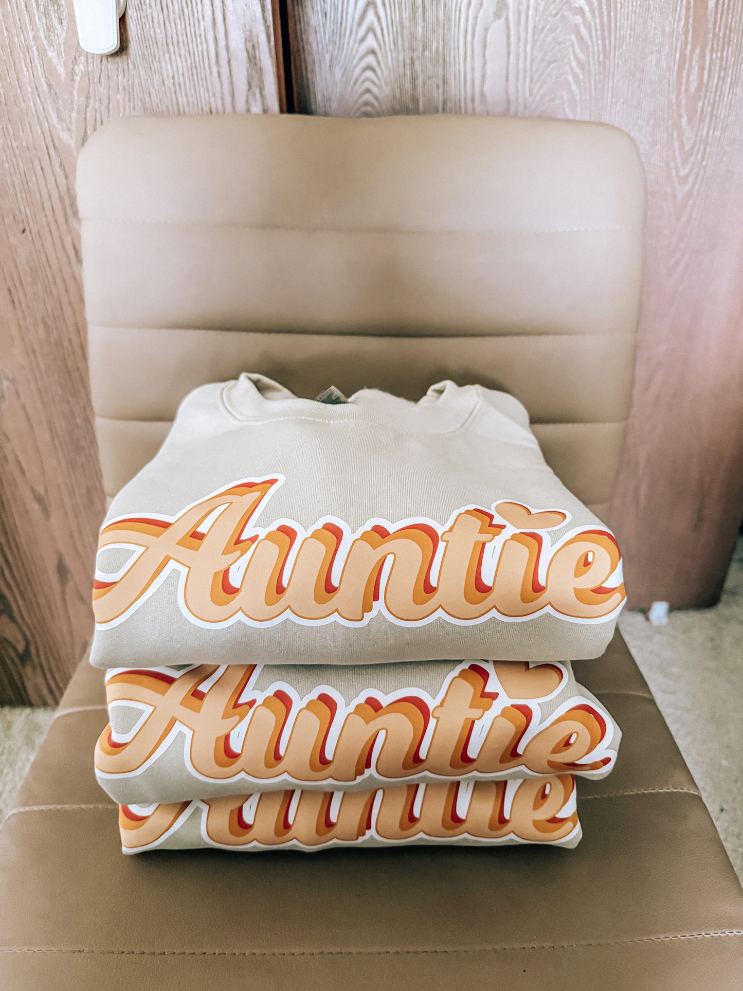 three sand sweatshirts with 'Auntie' printed on them, stacked on a beige chair.