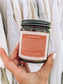 Hand holding a candle jar with a label against a neutral background