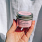 Hand holding a small pink jar with a silver lid against a light gray background